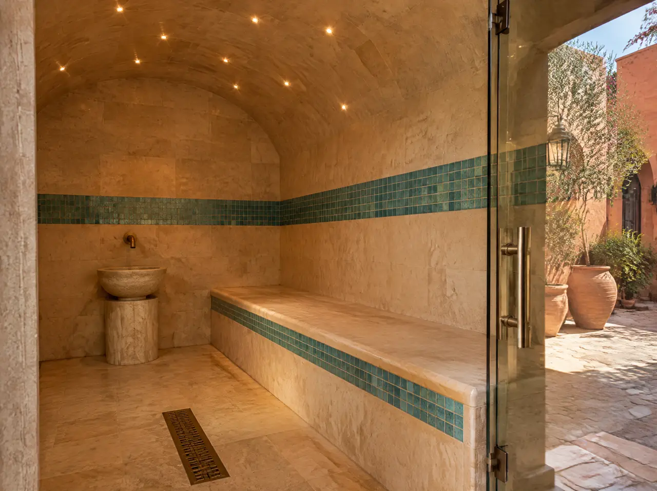 Interior seco de un baño de vapor de travertino crema y mosaico turquesa, con banca larga, techo abovedado con luces pequeñas y puerta de vidrio hacia un patio