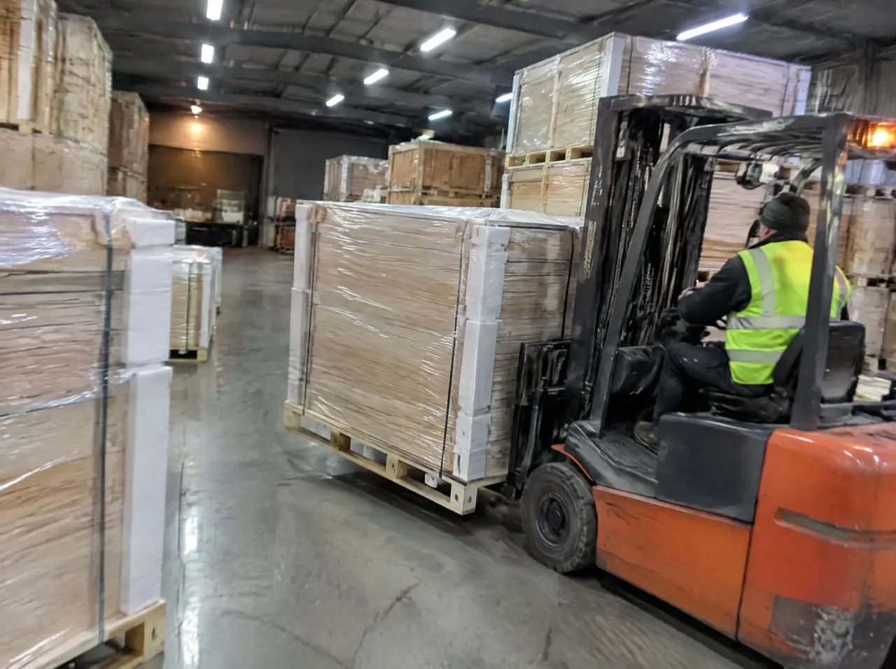 Cajas de madera contrachapada y tarimas con paneles de sauna envueltos en film y esquineros de espuma, con un operador de montacargas con chaleco reflectante