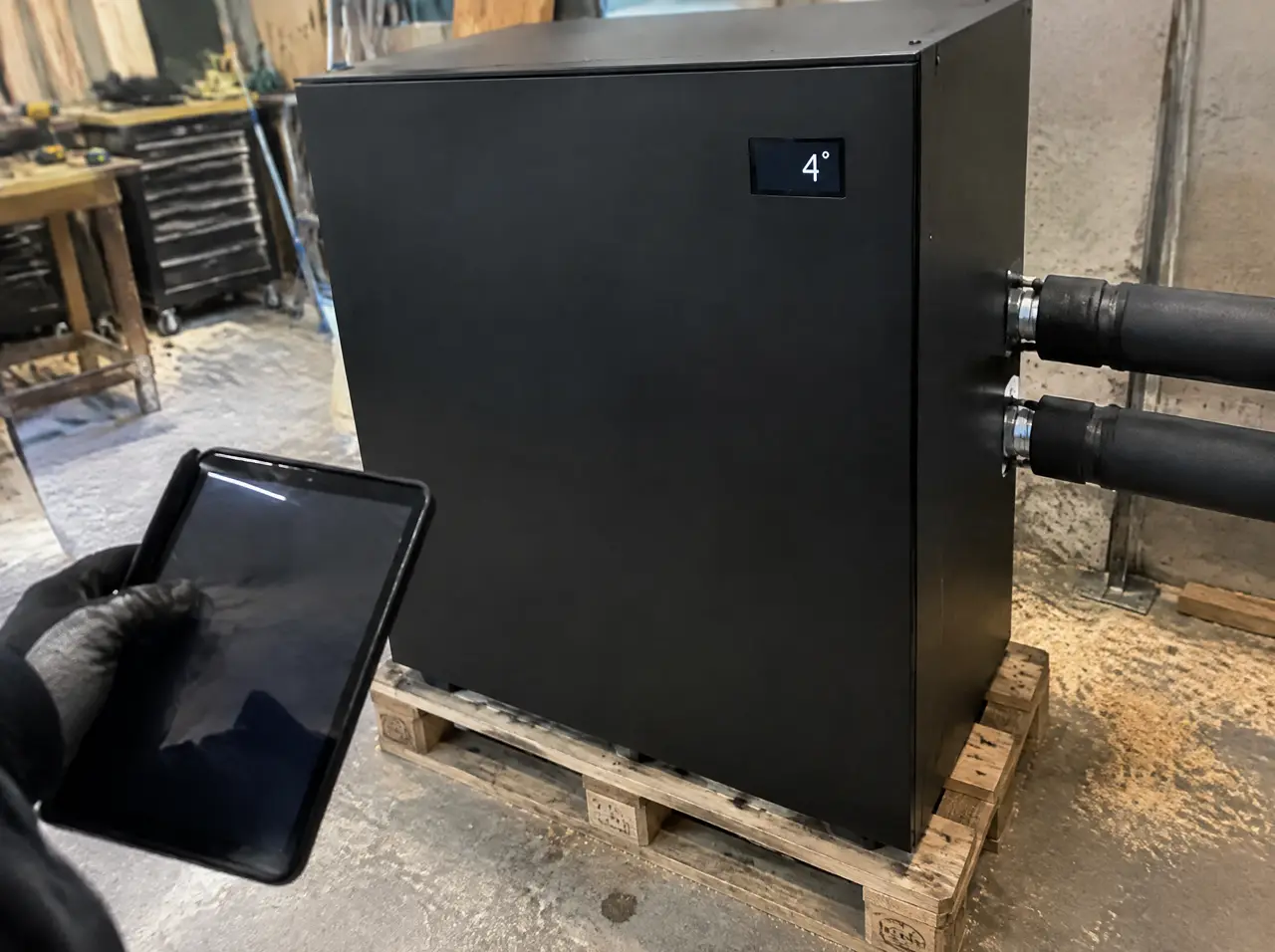 Caja negra lisa de un enfriador sobre una tarima en un taller, con dos tubos aislados saliendo de un costado y un panel pequeño que marca 4 grados