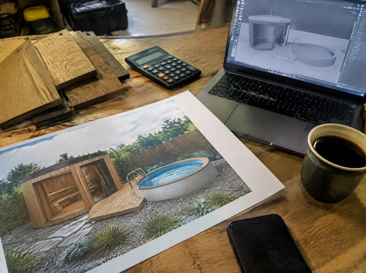 Escritorio con la impresión de un render 3D de un sauna y una piscina redonda, una calculadora, muestras de madera, una laptop con el modelo y un café