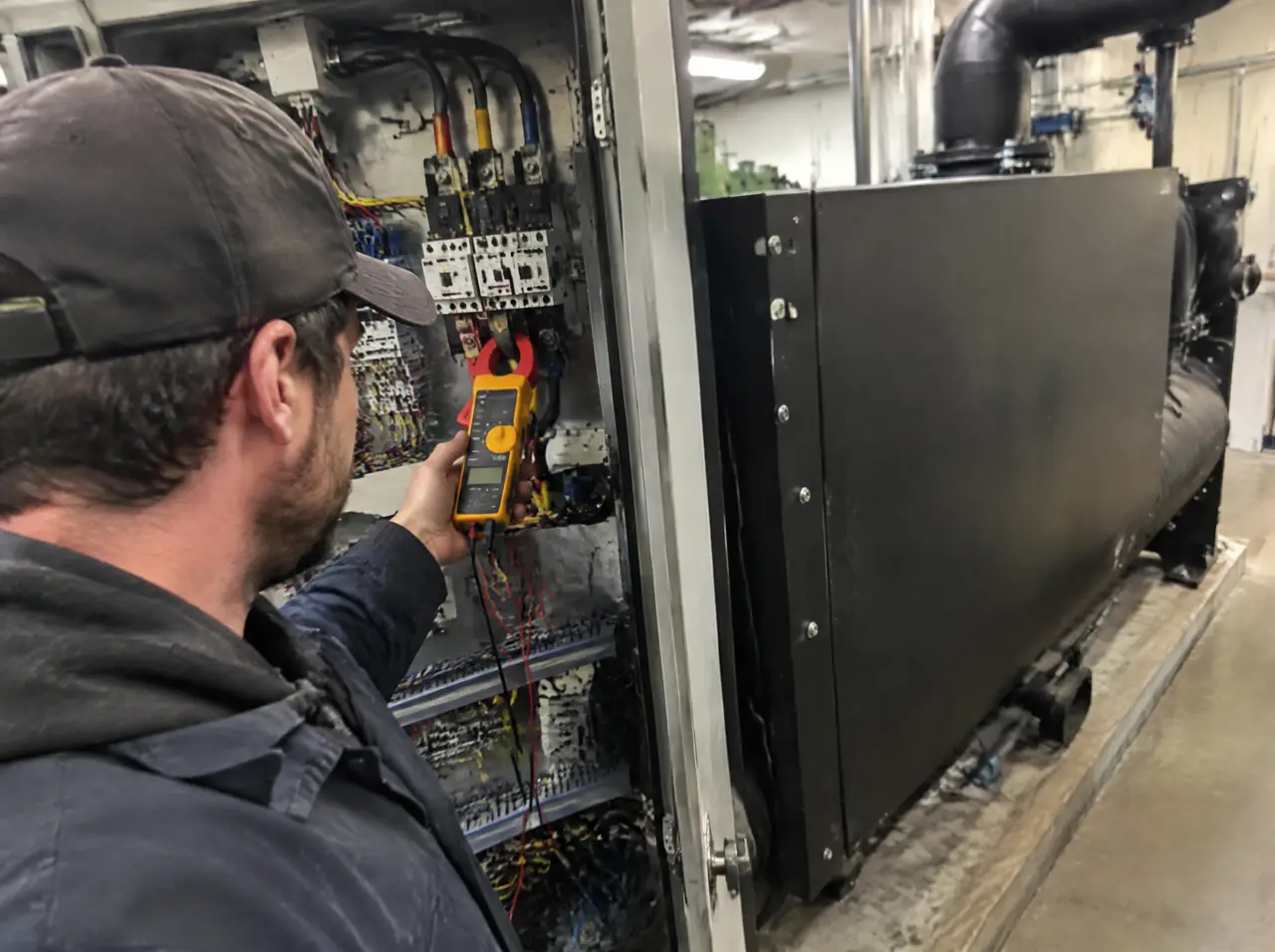 Técnico en un cuarto de máquinas midiendo con una pinza amperimétrica un cable dentro de un tablero abierto, junto a la caja negra lisa de un enfriador